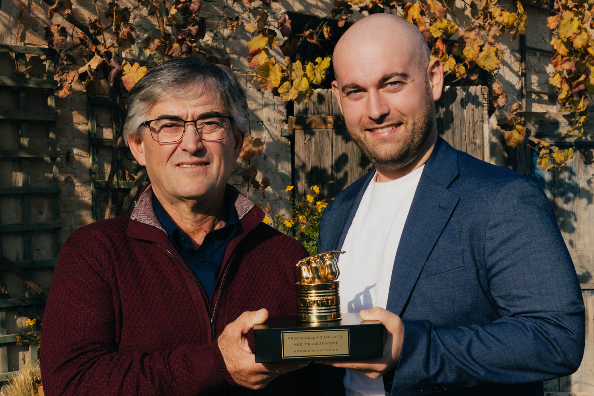 Two men holding the Pomodorino d'Oro trophy in front of autumn vines