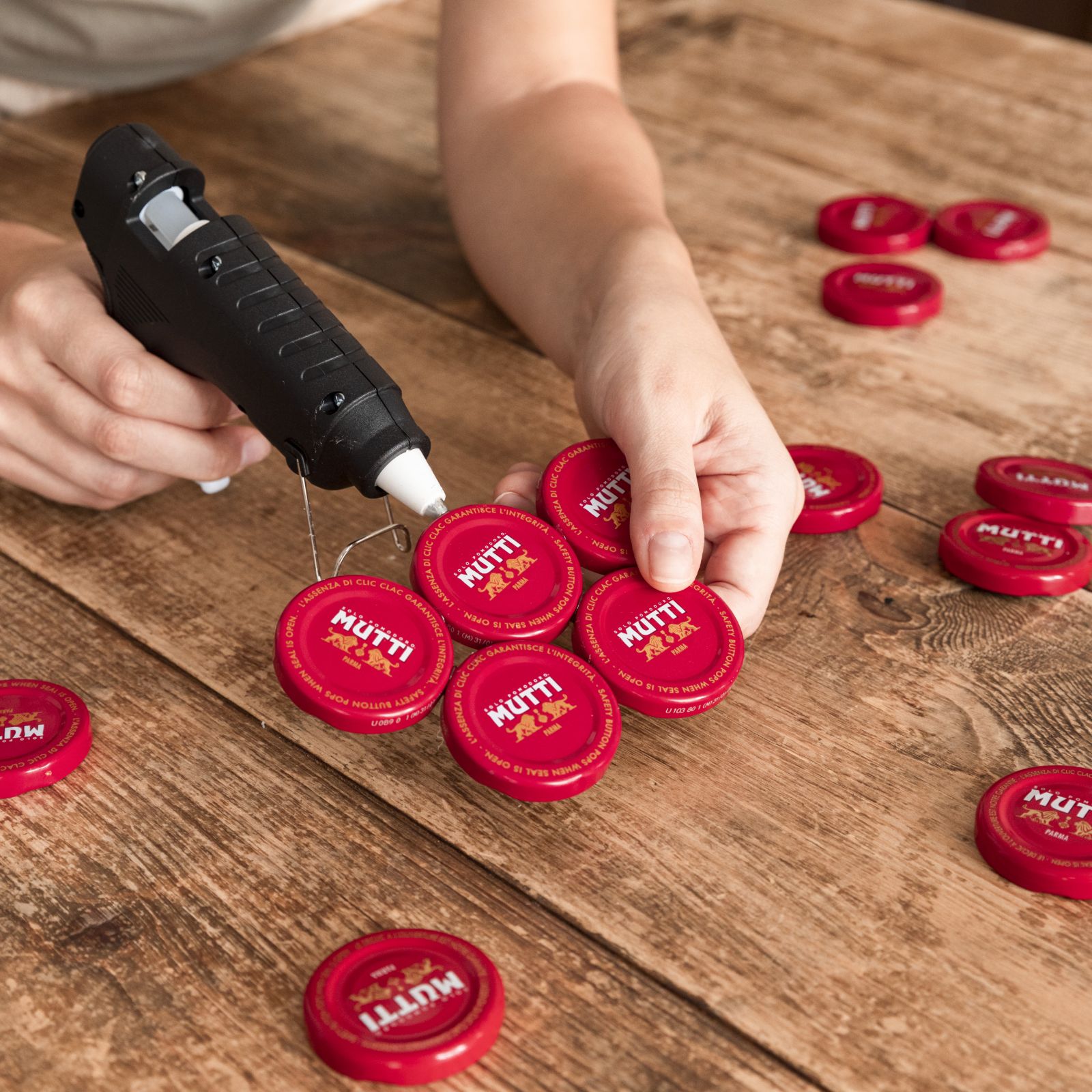 Hands hot-gluing red Mutti bottle caps together to form a coaster