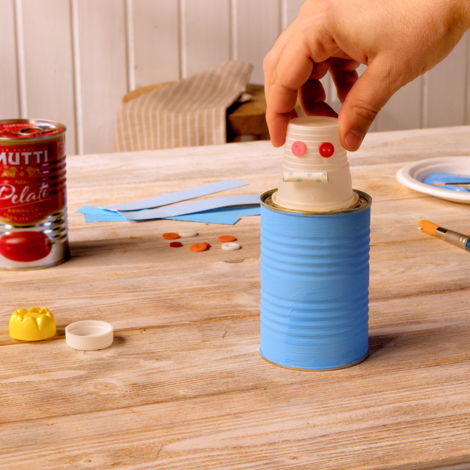 Hand placing a white cup head on a blue-painted Mutti can to build a toy robot