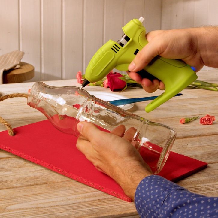 Hands hot-gluing a glass bottle onto a red canvas panel for a flower display