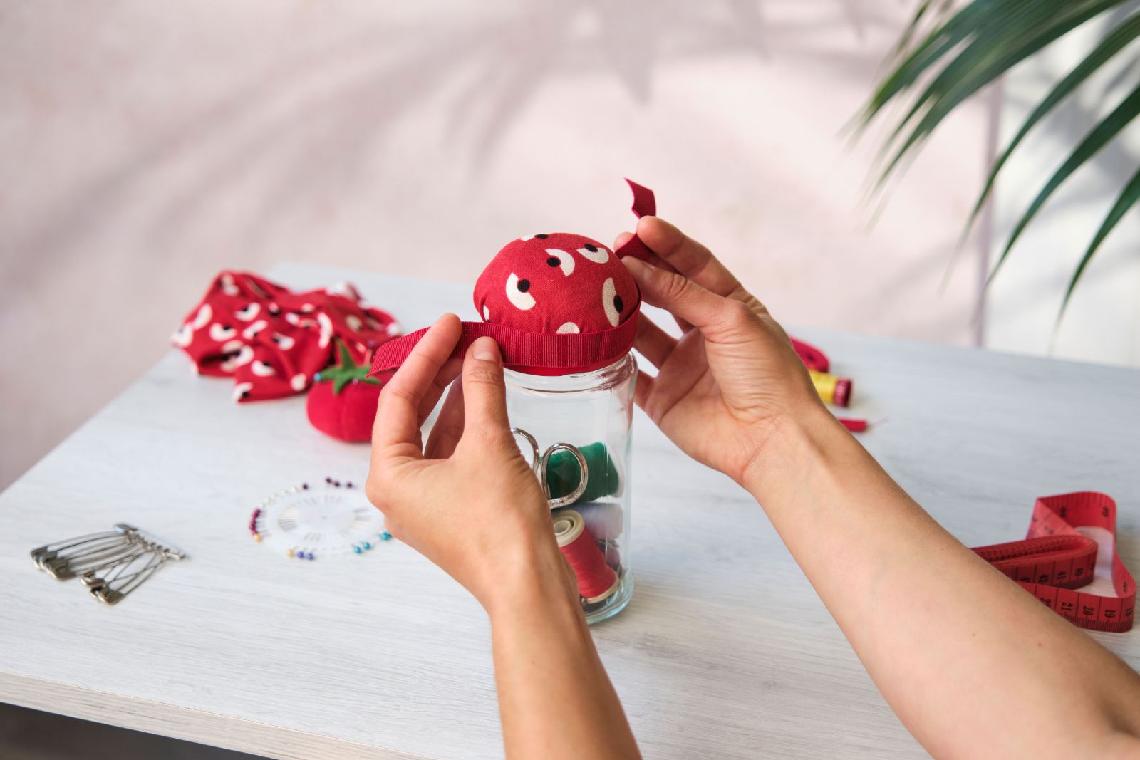 Attaching a red ribbon to a finished tomato-shaped pincushion on a jar